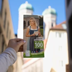 Der CityTourCard München Flyer wird von einer Frauenhand festgahlten. Im HIntergrund erkennt man die Türme der Frauenkirche in München
