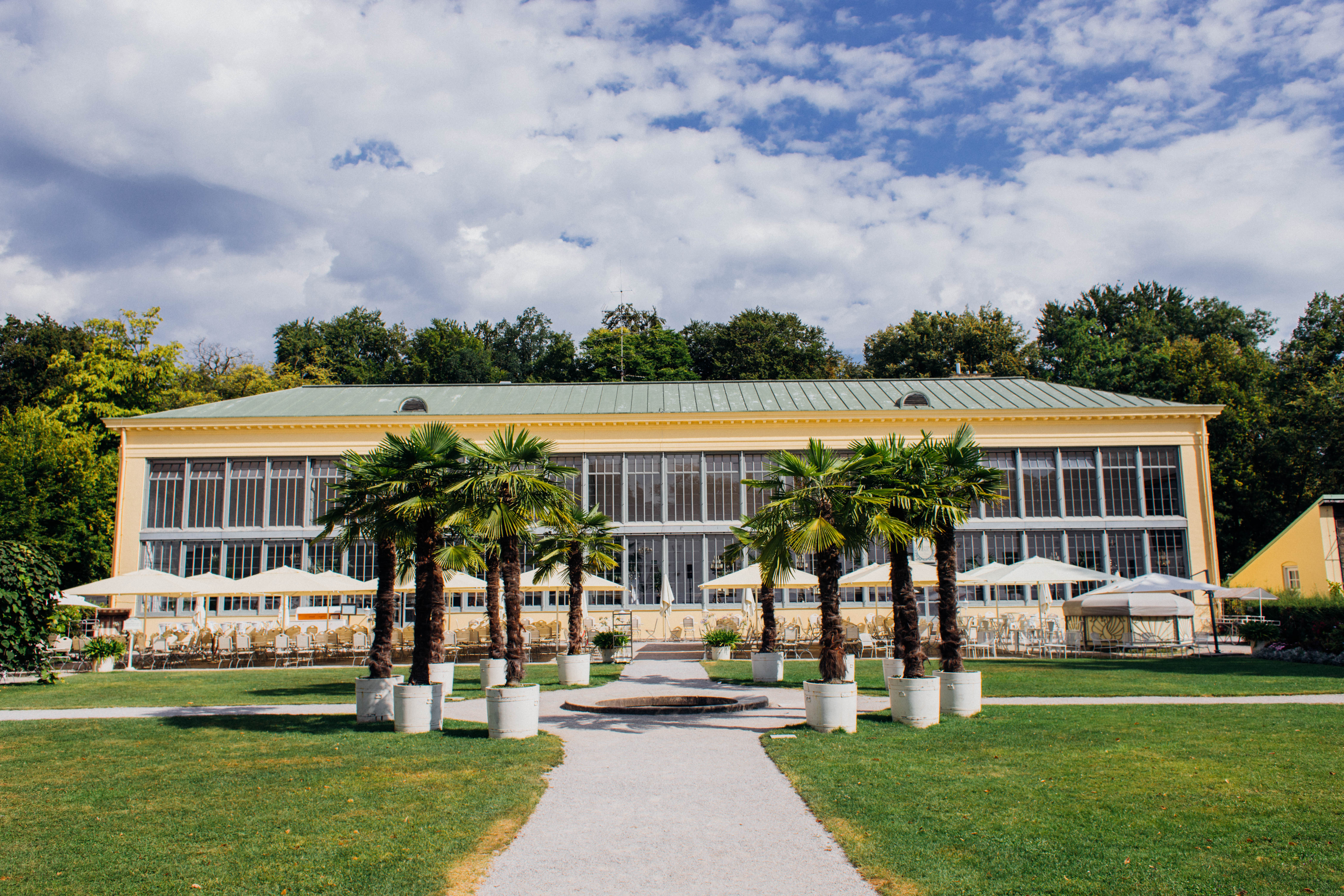 Das Schlosscafe Palmenhaus in Nymphenburg von außen. Es geht ein gerader Weg zum Haus mit Palmen an der Seite. Das Haus sieht aus wie ein Gewächshaus. Draußen stehen Tische, Stühle und Sonnenschirme