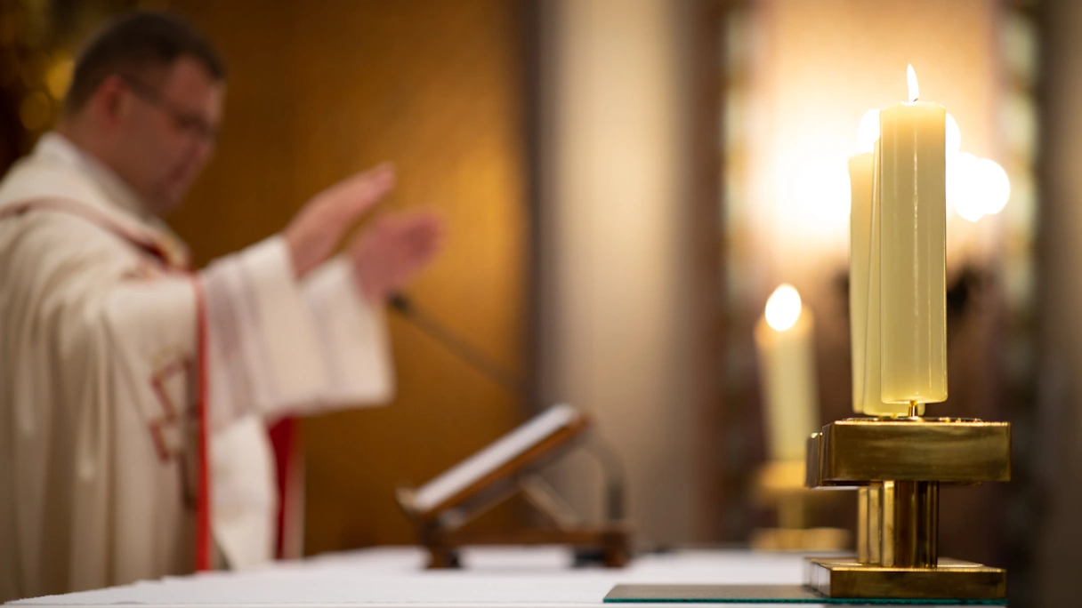 A priest prays with his hands raised, an open book lies in front of him. White candles are lit around him