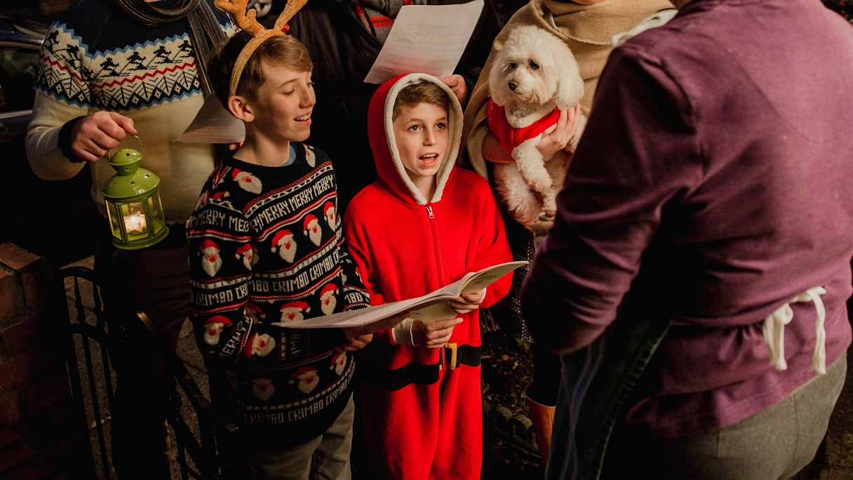 A family with two children and a dog are standing outside the front door of an elderly lady singing Christmas carols. They are wearing Christmas clothes