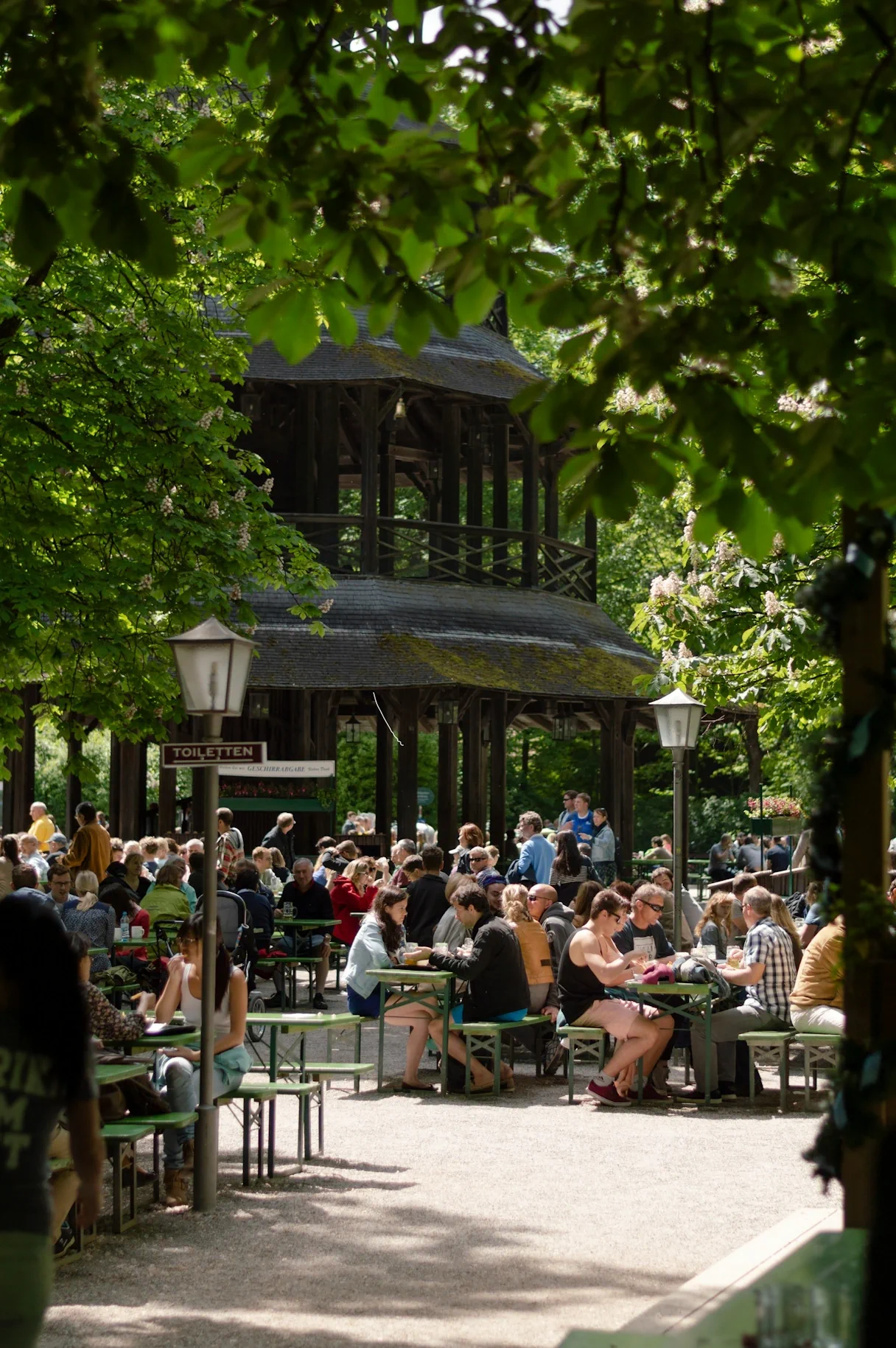 The beer garden at the Chinese Tower in the English Garden. There are beer benches and tables, most of the seats are taken, people are sitting, talking and drinking