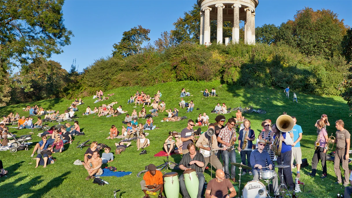 Englischer Garten in München, Wiese vor dem Monopteros, Menschen sitzen im Gras und genießen die Sonne. Am rechten Rand steht eine Musikgruppe mit verschiedenen Instrumenten