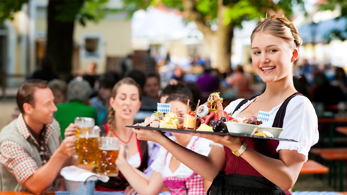 Biergarten in München, Kellnerin trägt ein volles Tablett mit einer Brotzeit, im Hintergrund sitzen Gäste, die mit einer Maß Bier anstoßen