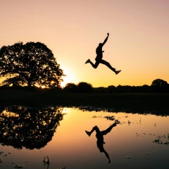 Sonnenuntergang an einem See. Ein Mann Springt in die Luft, daneben steht ein großer Baum. Der Mann und der Baum spiegeln sich im Wasser vom See