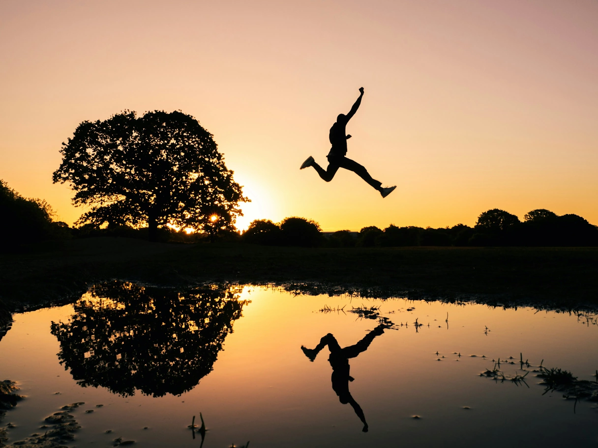 Sonnenuntergang an einem See. Ein Mann Springt in die Luft, daneben steht ein großer Baum. Der Mann und der Baum spiegeln sich im Wasser vom See