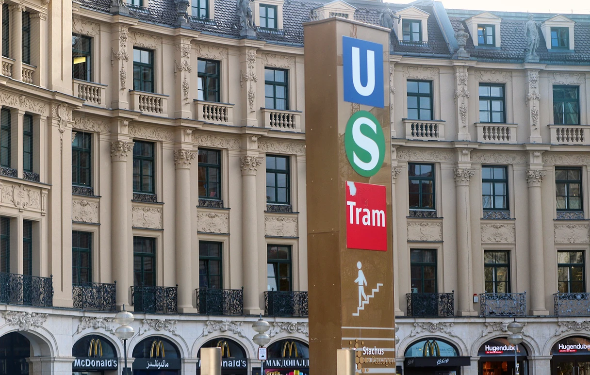 Karlsplatz, Stachus, Stele mit dem U-Bahn, S-Bahn und Tram-Zeichen, im Hintergrund eine Häuserzeile