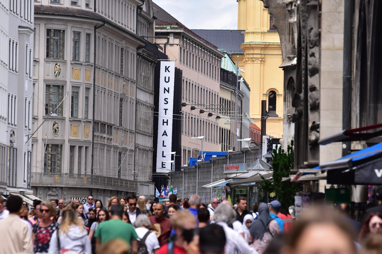 Straße in München die viel belaufen ist. Links und rechts sind Gebäude zu sehen. An einem Gebäude hängt ein großer Banner mit der Aufschrift "Kunsthalle"