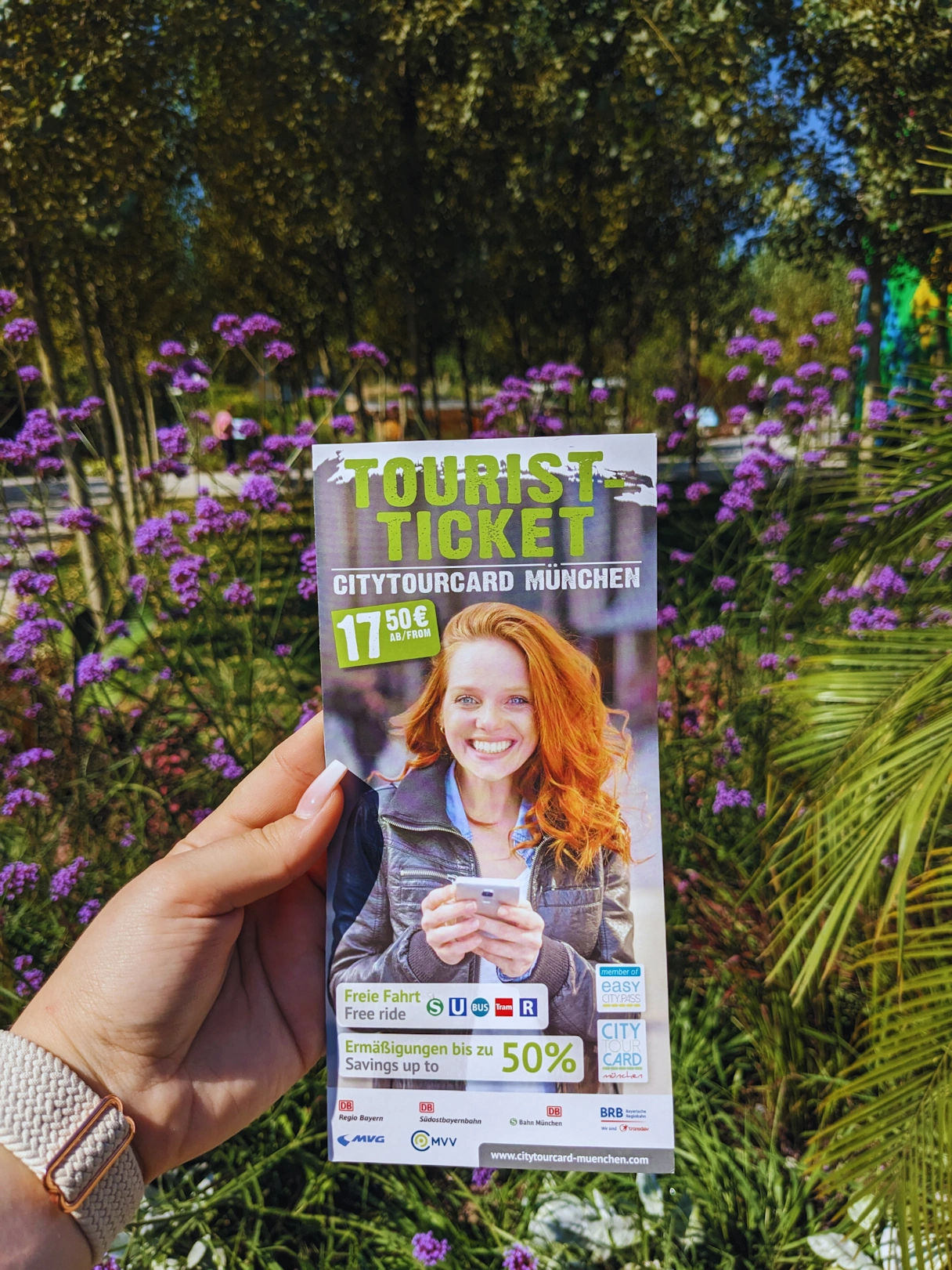 The CityTourCard Munich Flyer in one hand. There are green and blue/purple plants in the background