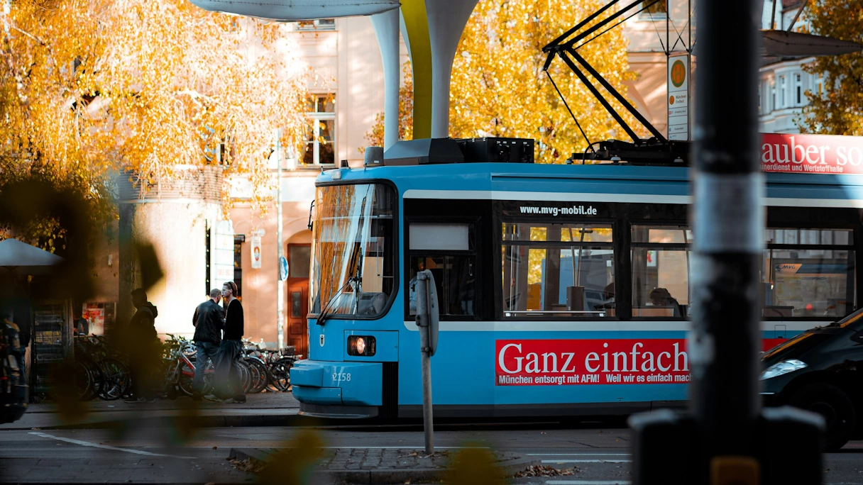 Die Tram in München an einer Haltestelle im Herbst. Auf der Tram befindet sich rote Bannerwerbung