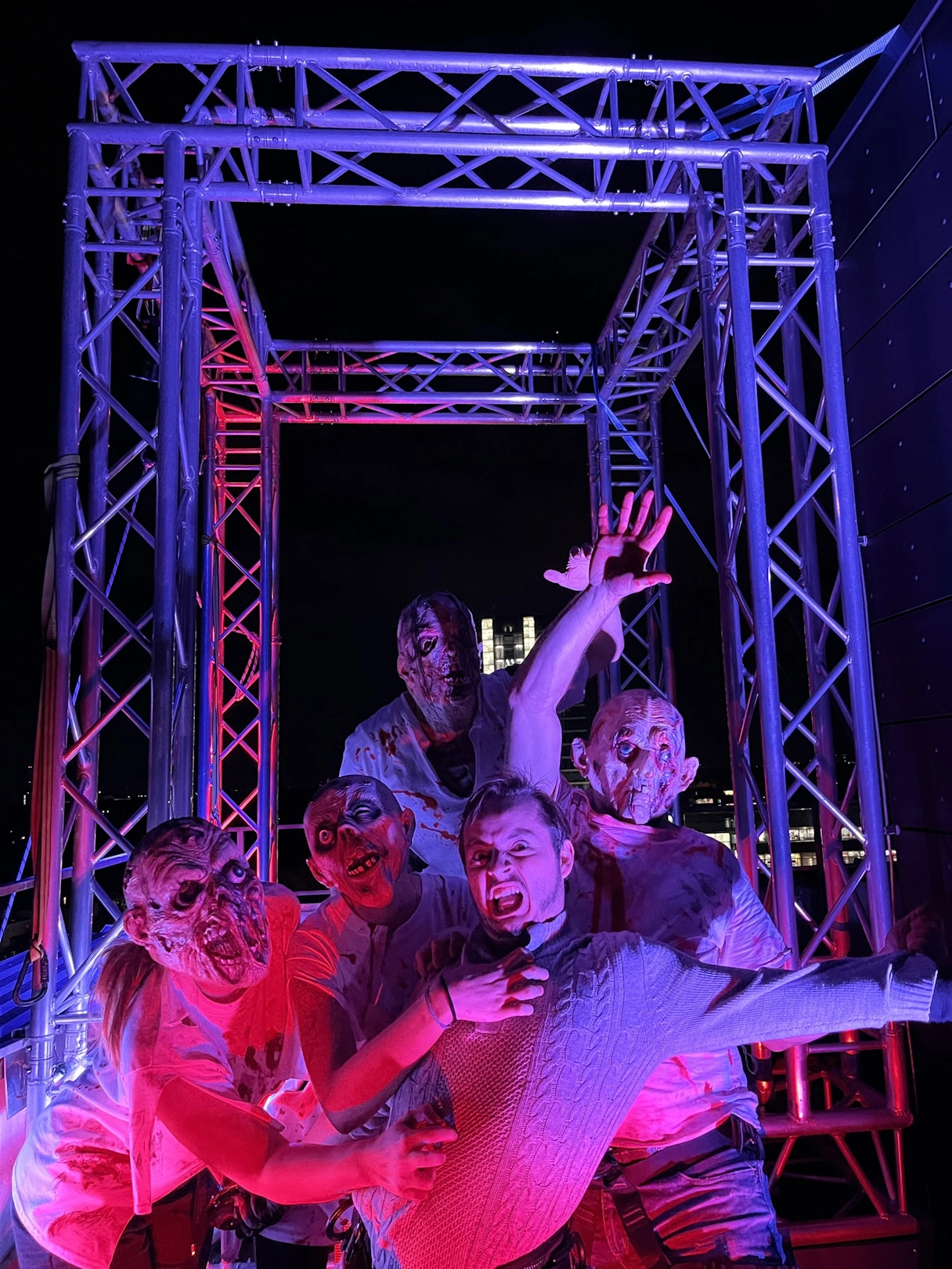 4 people dressed up as monsters and a frightened looking person in the middle. It is dark and they are illuminated by colored light