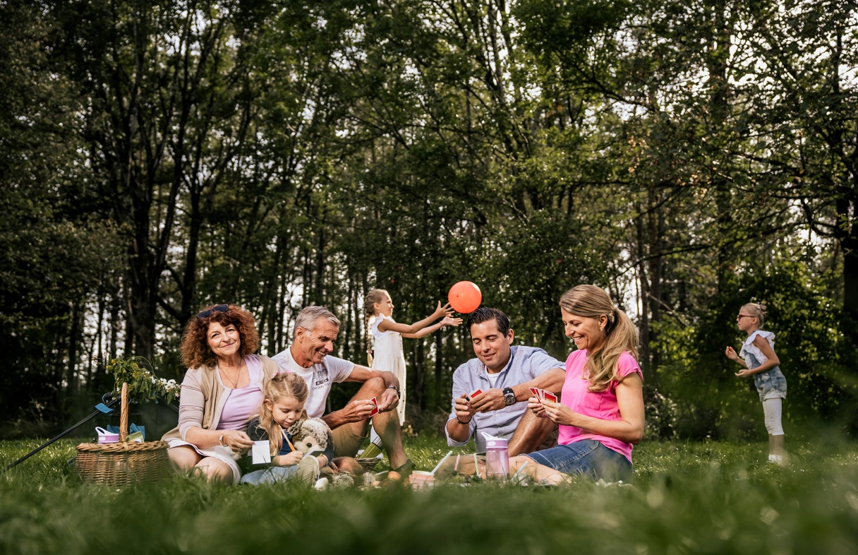 Familie sitzt fröhlich auf der Wiese und macht ein Picknick. Kinder spielen im Hintergrund