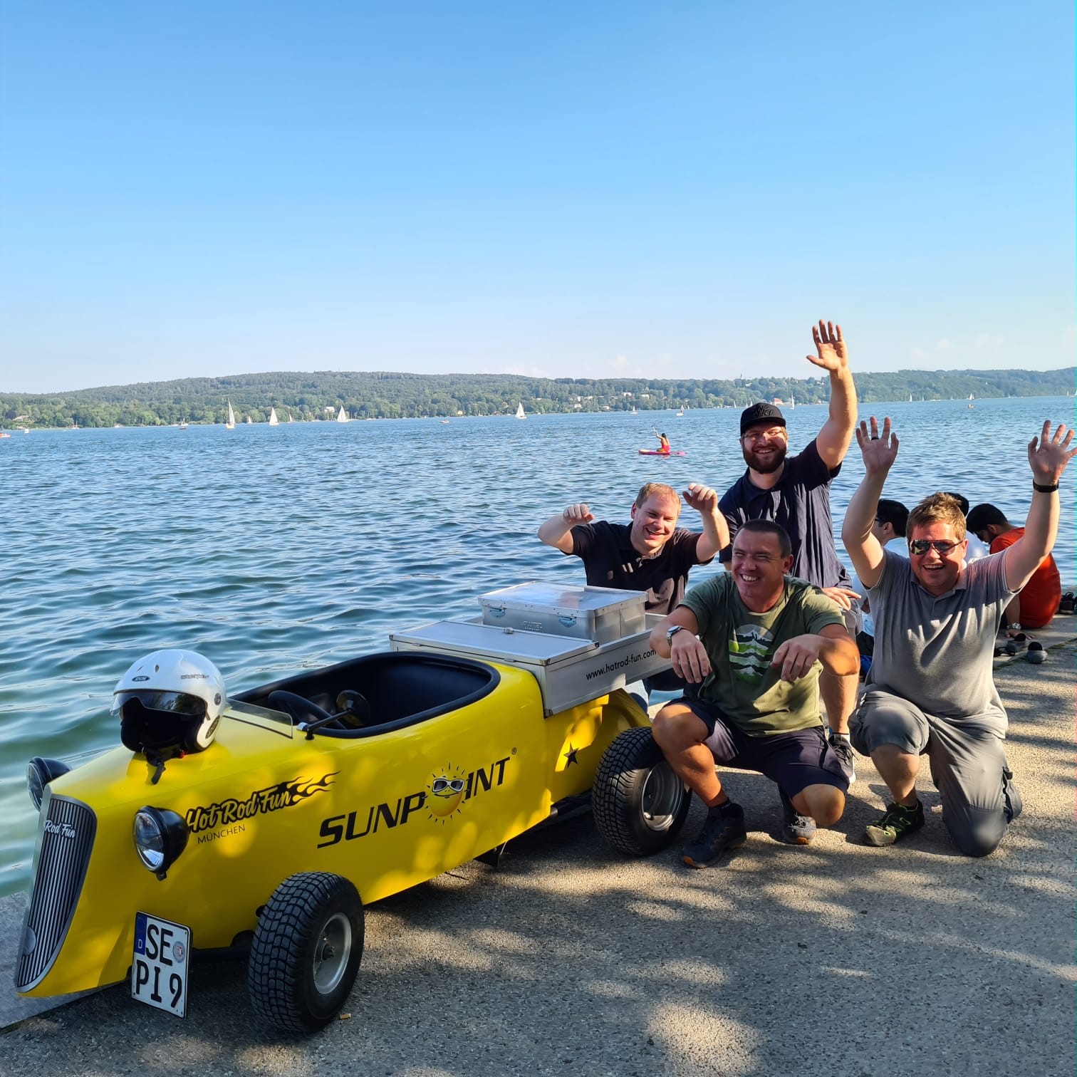 Ein Hot Rod Kart am Starnberger See, daneben sitzt die Gruppe und winkt und lächelt fröhlich.