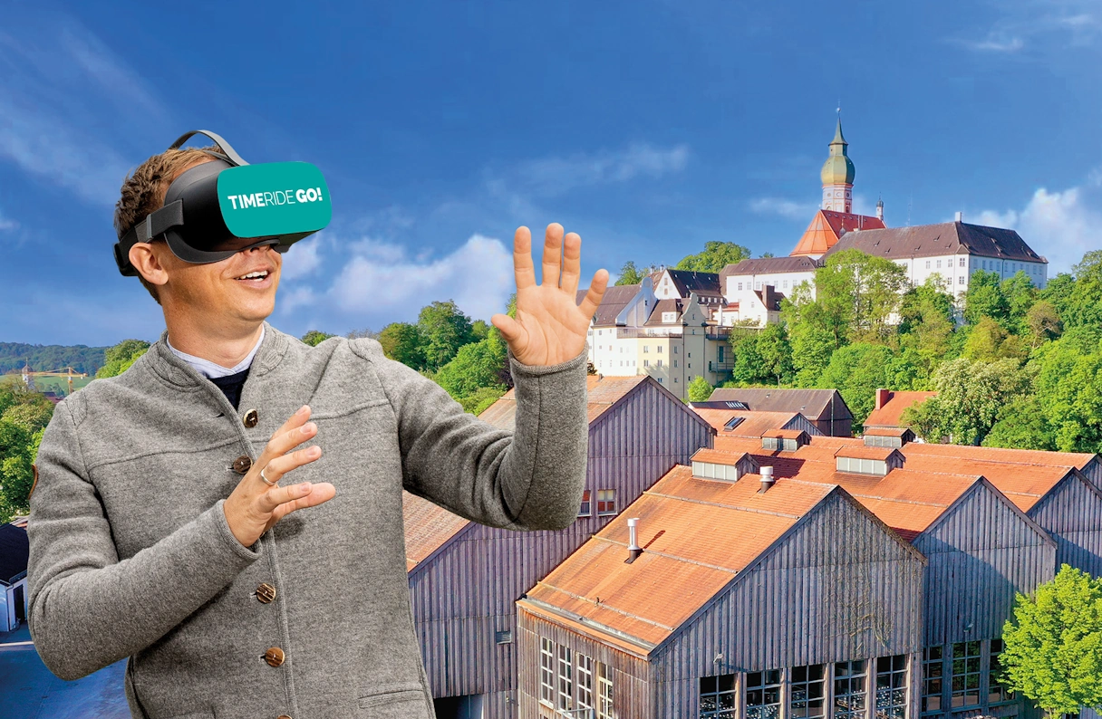 ein Mann mit VR Brille. Im Hintergrund sind Häuser, Bäume und das Kloster Andechs zu sehen