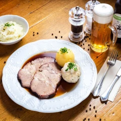 Schweinebraten mit Klößen serviert. Daneben steht eine Portion Krautsalat und ein Glas Bier