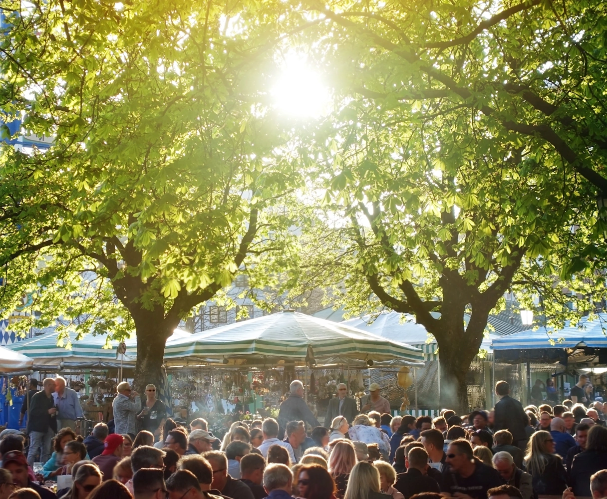 Bieragrten am Viktualienmarkt von vielen Menschen besucht. Durch die Baumkronen scheint die Sonne