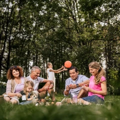 Familie sitzt gemeinsam auf einer Wiese. Die Kinder spielen und alle sehen fröhlich aus