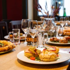 angerichtetes Essen in den Münchner Stubn. Bayerisches Essen sowie Pizzen. Außerdem sind leere Weingläser zu sehen.