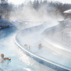 Der Außenpool-Strömungskanal in den Chiemgau Thermen Bad Endorf. Dre Menschen schwimmen darin im Winter, während draußen Schnee liegt