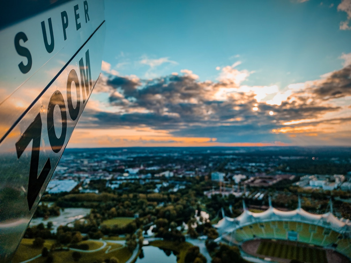 Der Olympiapark von oben fotografiert. Im Vordergrund ist ein Super Zoom Fernglas zu sehen, im Hintergrund das Olympiastadion, teils bewölkter Himmel und die Skyline