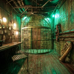 Raum bei Escape Game München. Dunkle Steinwand, dunkler Holzboden, Rohre an der Decke und an der Wand. In der Mitte ein Käfig mit einer Tür in der Wand. links hängen Spiegel, Bilder und ein Tisch mit einer Nähmaschine.