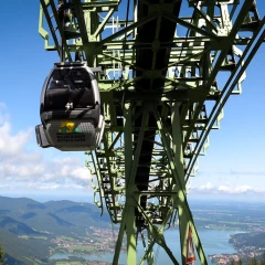 Die Wallbergbahn. Ein Gondel befindet sich weit oben, im Hintergund sieht man Berge, Wälder und den Tegernsee