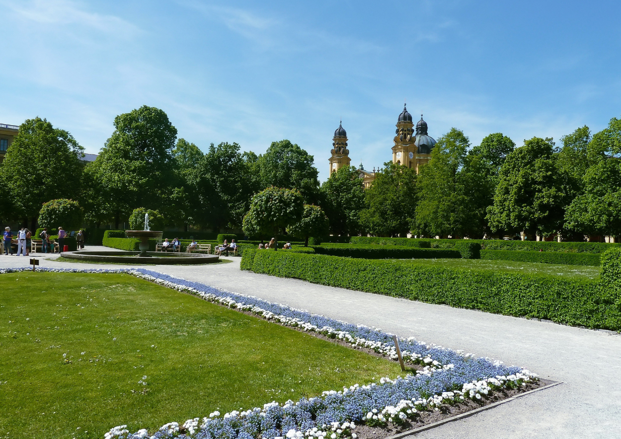 The Munich Hofgarten in summer. Green meadow, bushes, trees, a small fountain in the middle and the Theatinerkirche church further back in the background