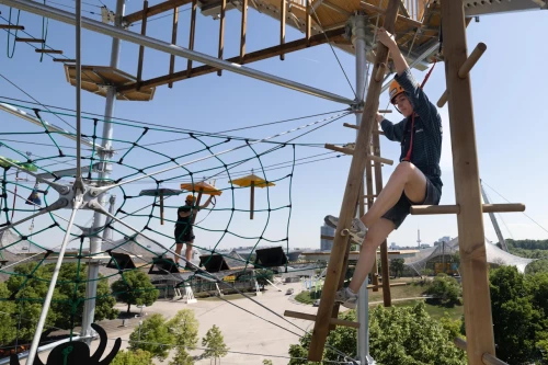 Crystal Tower/Adventure Tower in Munich's Olympic Park. People are climbing ropes, nets, and beams while wearing safety gear.