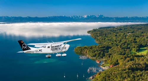The Alpen Air Cessna aircraft flies over Lake Starnberg. The water is blue, there are only a few clouds in the sky. Mountains can be seen in the background
