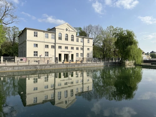 The Alpine Museum from the outside. A white, medium-sized building with the Isar river flowing in front of it, trees on the left and right.
