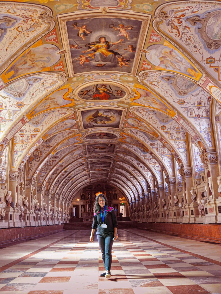 Guided tour of the residence and Diana stands in the center of the picture. Richly decorated ornaments on the ceiling and checkerboard pattern on the floor.