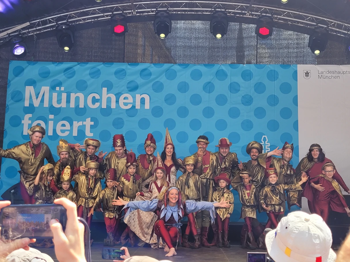 A group of costumed people are lined up on a stage in front of a blue wall that reads “Munich celebrates”. The audience takes photos.