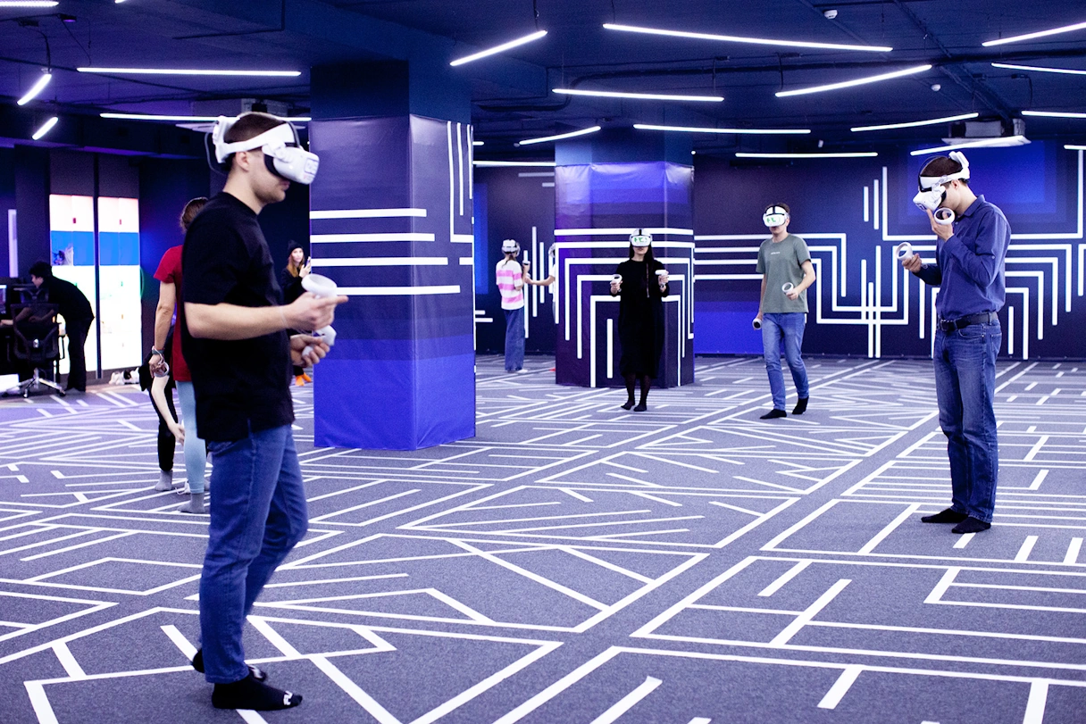 VR Spielehalle. Zu sehen sind Menschen in einem großen Raum, die VR Brillen tragen und Controller in der Hand halten. Auf dem blauen Teppich und an der blauen Wand sind weiße Linien.