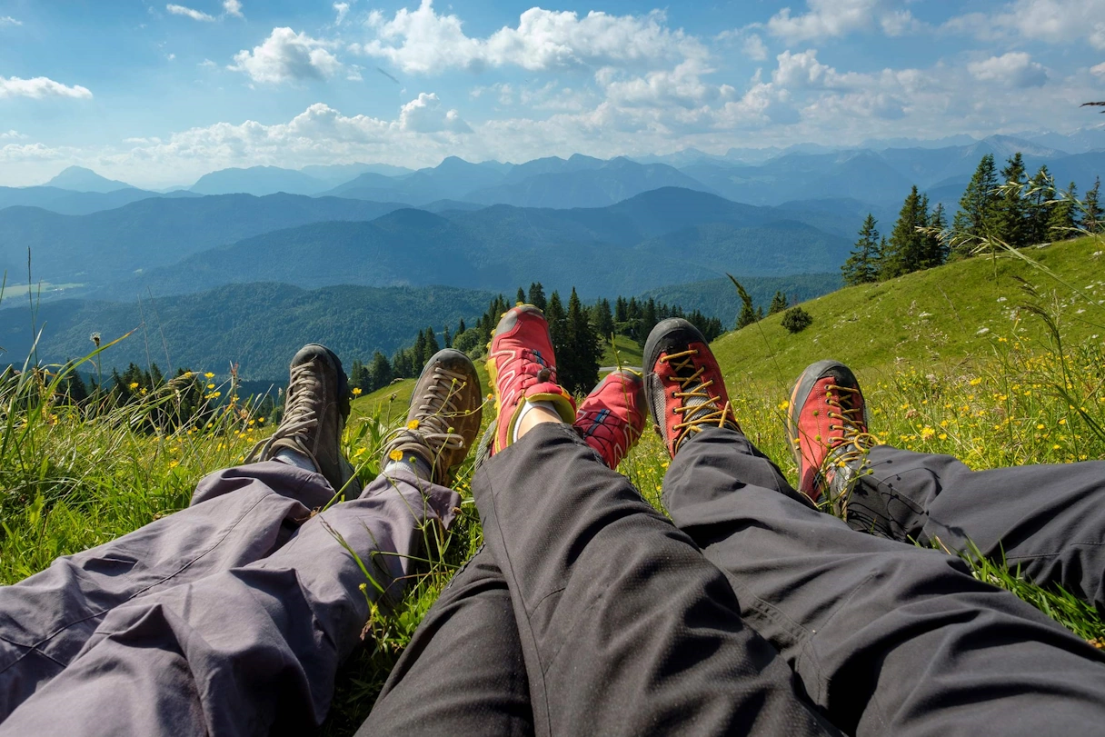 3 pairs of legs in hiking pants and hiking boots lying on the meadow + a view with mountains and landscape in the background