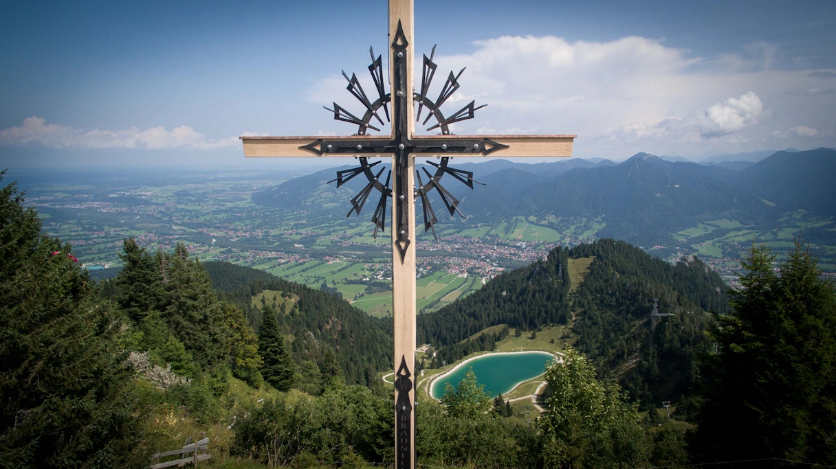 Das Brauneck Gipfelkreuz, im Hintergrund sind Berge, Landschaft und ein See zu erkennen