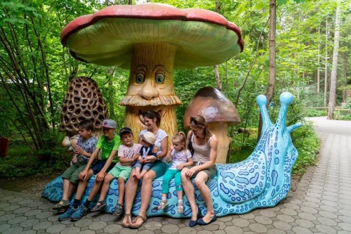Fairy tale forest Munich, big mushroom with hat and face stands on the roadside, in front of it a blue snail used as bench, family with several children sitting on it