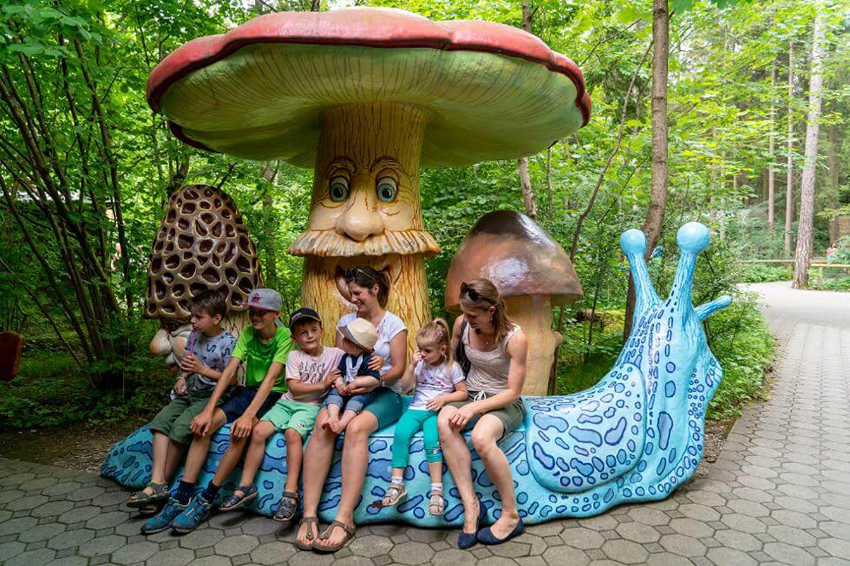 Märchenwald München, großer Pilz mit Hut und Gesicht steht am Wegesrand, davor eine blaue Schnecke, die als Bank benutzt wird, Familie mit mehreren Kindern sitzt drauf