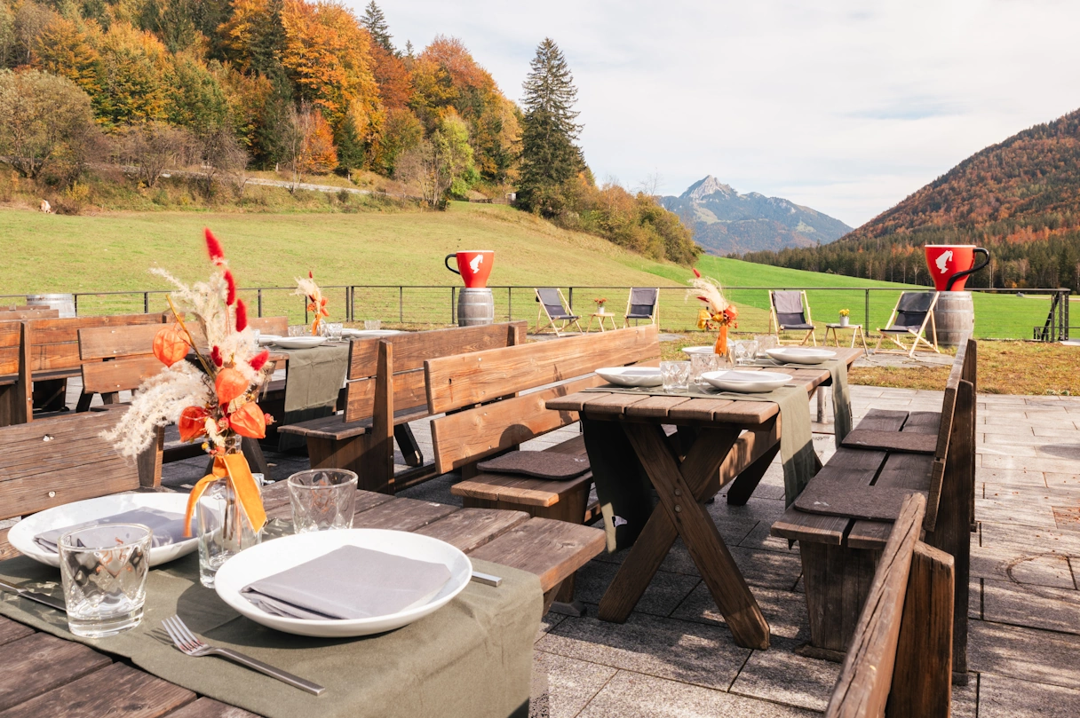 The outdoor area for guests of the GenussVerweilerei am Schliersee. Several wooden tables with wooden benches for approx. 6-8 people. Mountains and green meadows can be seen in the background.