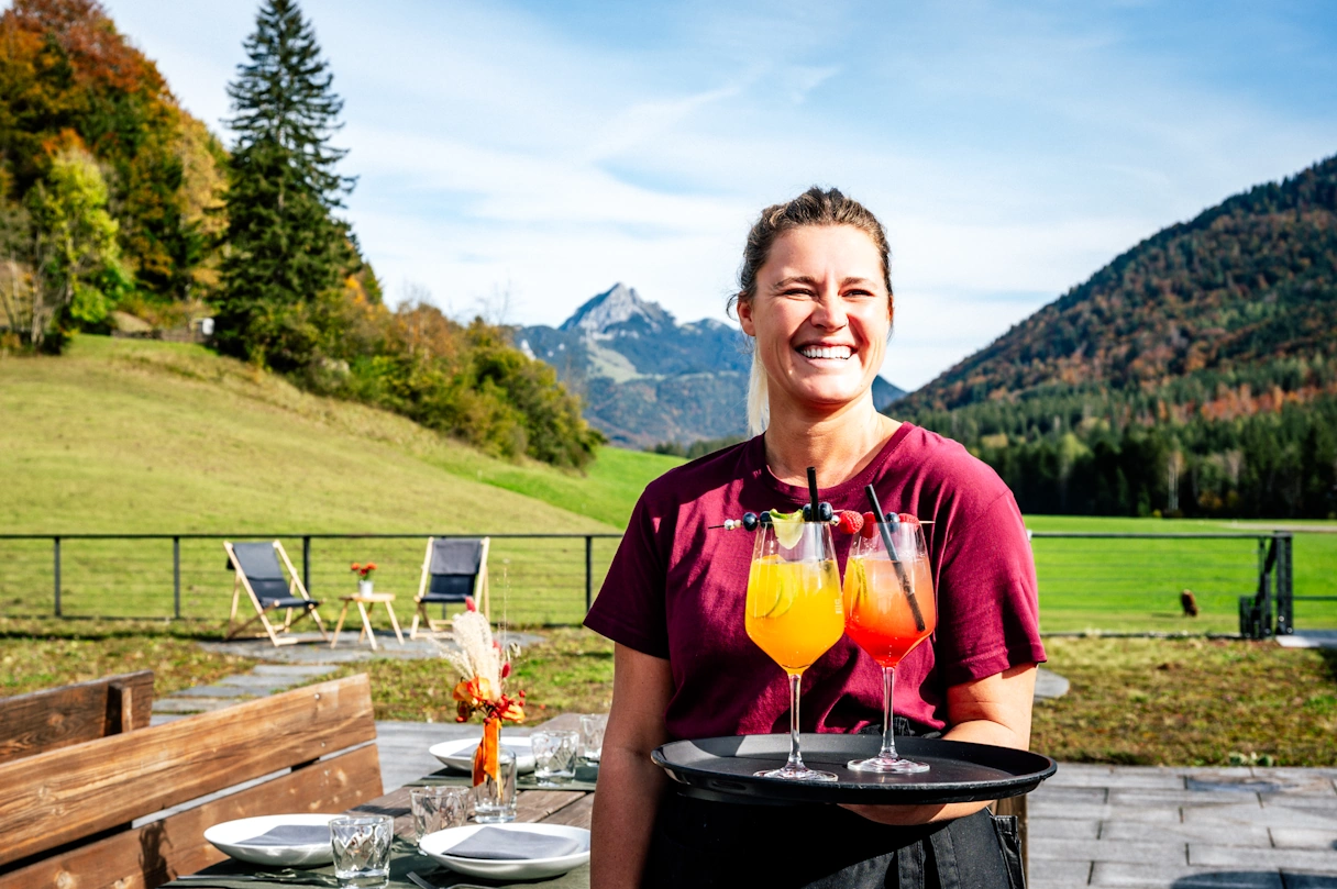 Waitress stands outside with a tray and laughs. On the tray are two wine glasses filled with an alcoholic, colorful drink. In the background are mountains and green meadows