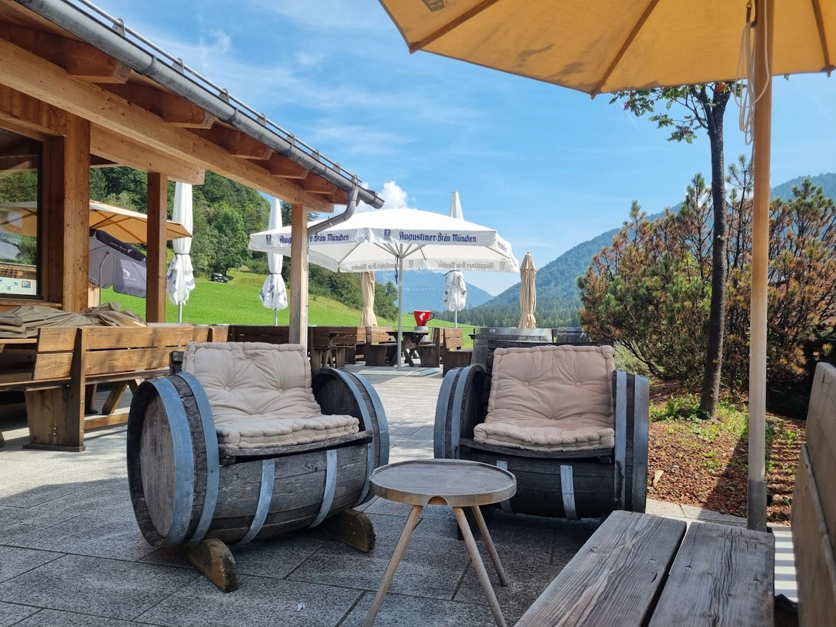 Outdoor area Genussverweilerei am Schliersee. Sunshades, small wooden tables and wooden barrels that have been converted into seating