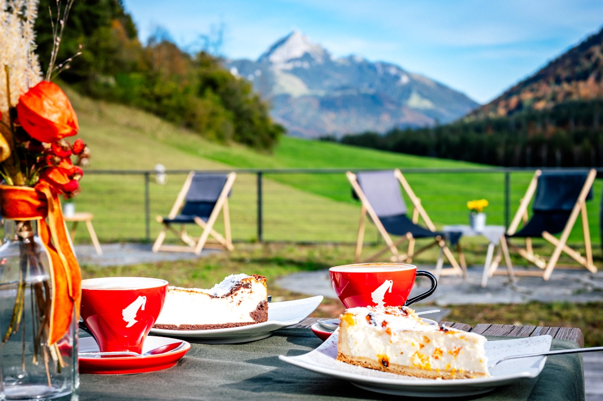 Two plates, each with a piece of cake, next to two cups of coffee and a vafe with flowers. In the background are mountains and a green meadow, as well as other seating areas