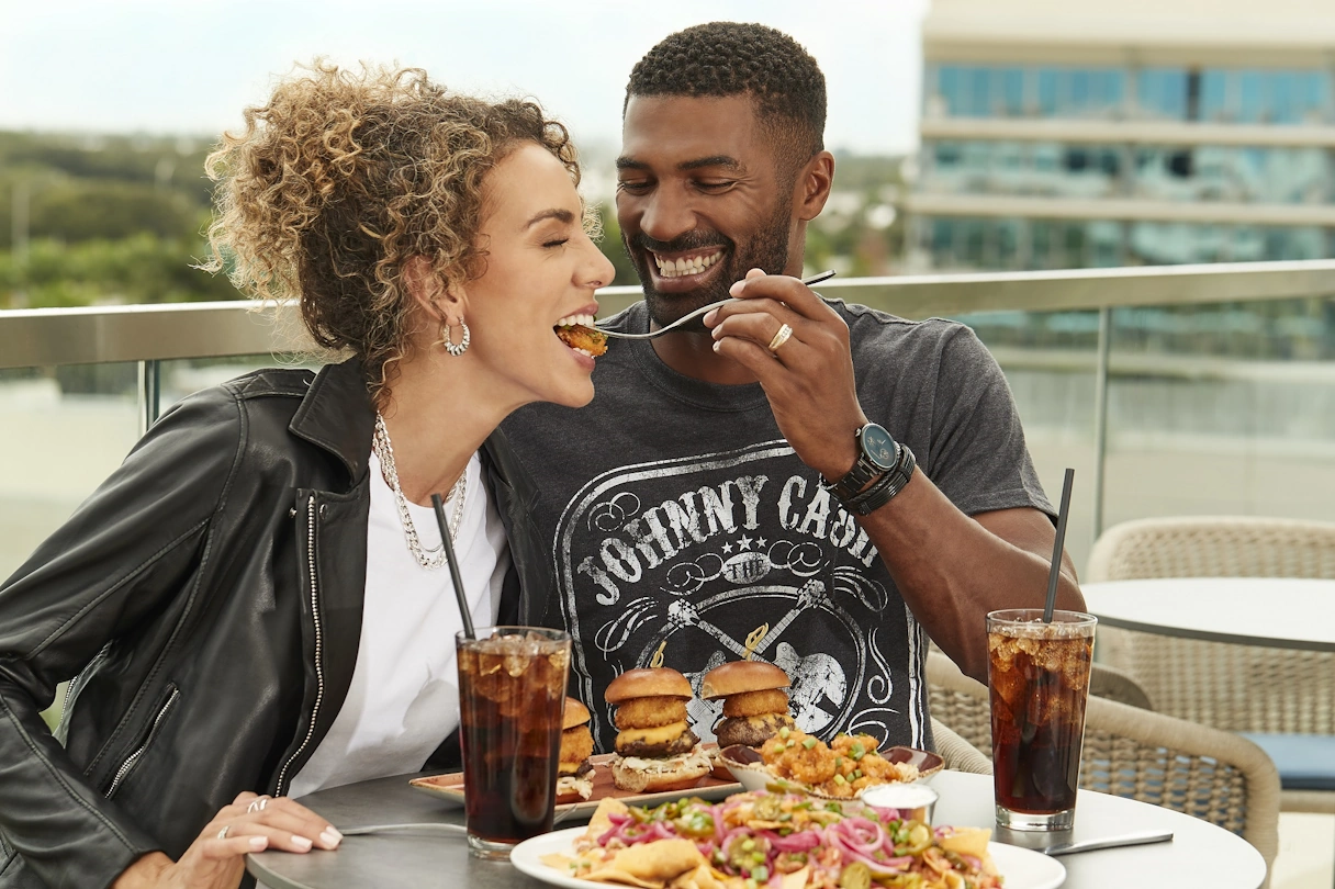 Tisch gedeckt mit Burgern und zwei Gläsern Cola, Mann schiebt einer Frau lachend die Gabel mit dem Essen zum Mund