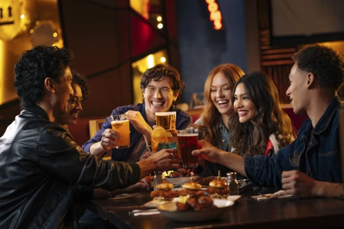 Three young men and three young women in the Hard Rock Cafe. There are burgers, chicken wings and other food on the tables. The group laughs and has drinks in hand for a toast