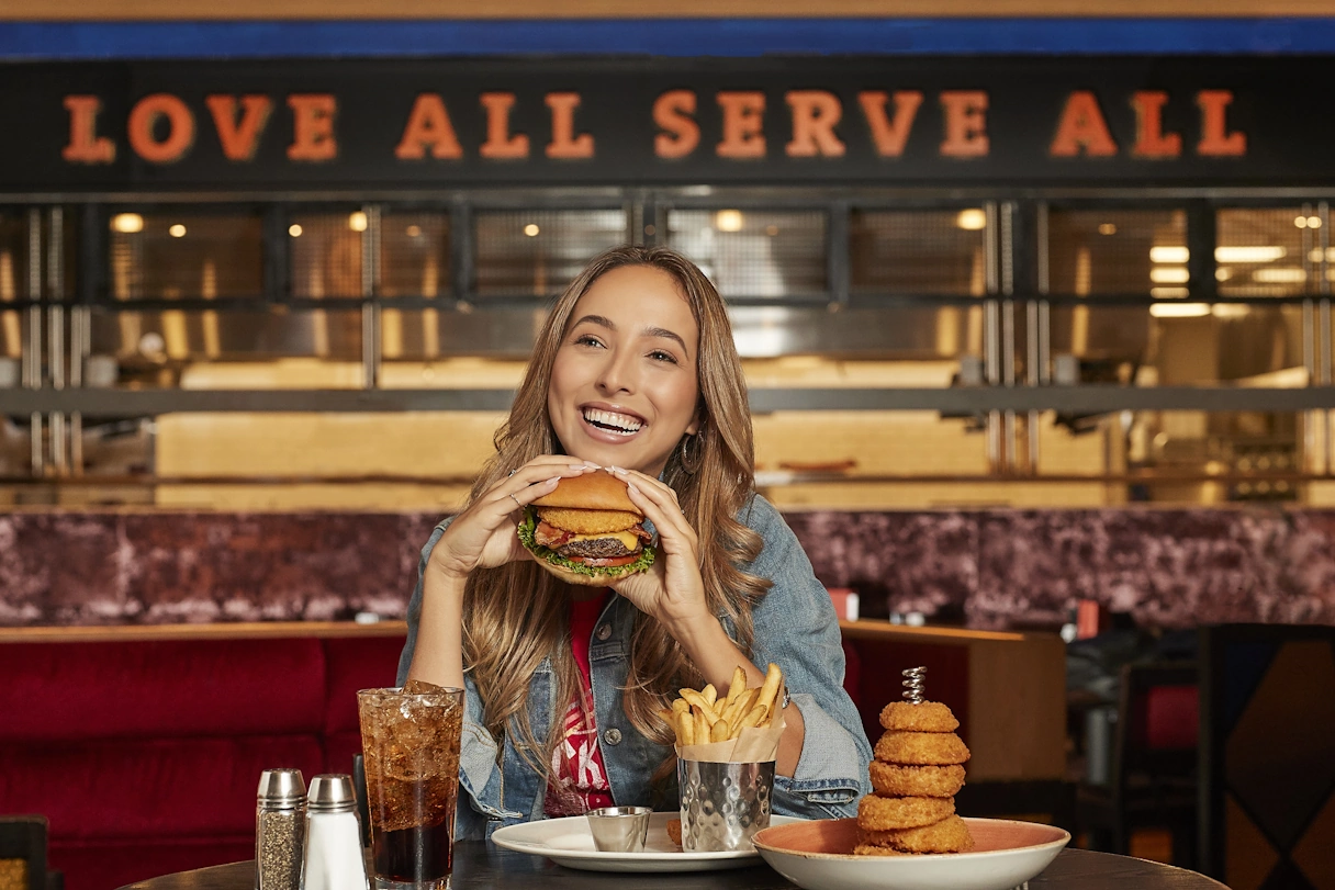 Frau im Hard Rock Cafe hält lachend einen burger in der Hand. Vor ihr stehen Getränk und Pommes