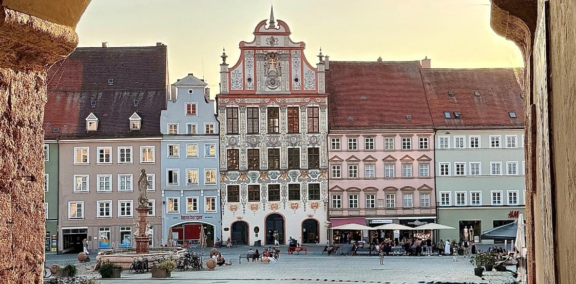 A view of the historic town hall in Landsberg am Lech. In front of it is the main square, featuring a fountain and places to sit. The building facades are colorful and create a cozy atmosphere.