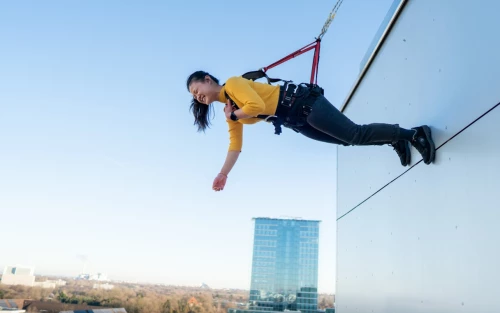 A young woman doing house running in Munich. She is hanging from the exterior of a building with a safety harness and laughing.