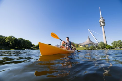 A young man sits in a kayak and paddles across the Olympic Lake in Munich. In the background, you can see the Olympic Tower and part of the stadium.