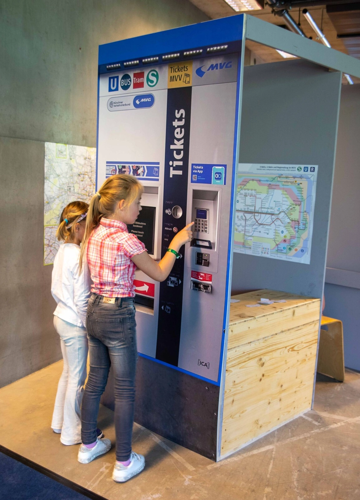 2 girls stand at a sample ticket machine made of cardboard