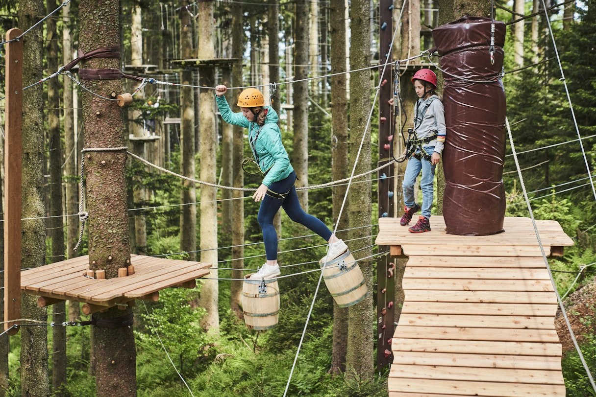 Bereich im Kletterwald Blomberg. Eine junge Frau balanciert über zwei hölzerne Fässer, die in der Luft hängen. Hinter ihr wartet ein Kind