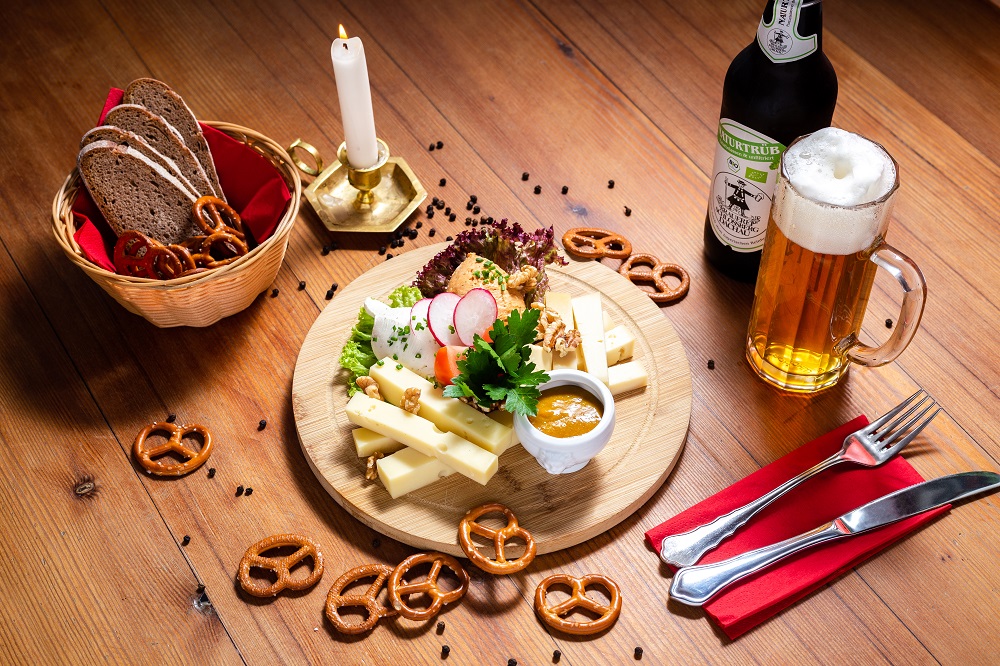 Käse und Gekmüse auf einem runden Brett mit kleinen Salzbrezeln als Deko, einem Bier, einer Kerze und einem Korb mit Brot