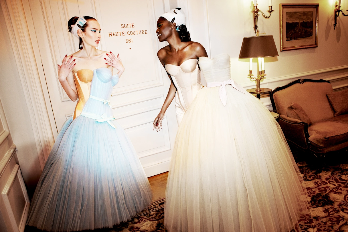 Two women are wearing a haute court dress with a large tulle skirt and colorful make-up. They are standing in a hotel room